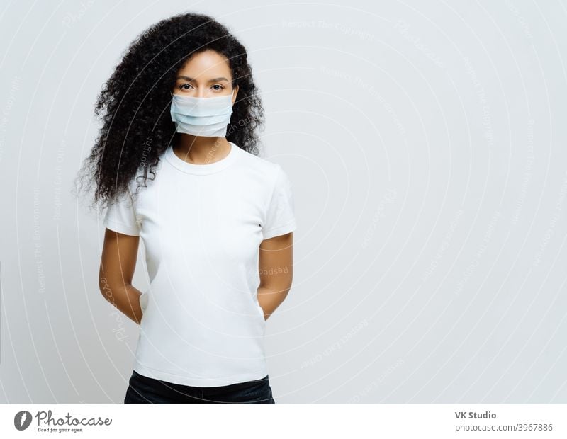 Half length shot of Afro American woman being on self isolation or quarantine, wears medical mask during coronavirus outbreak, listens news, tries not to spread virus, stays at home for long period