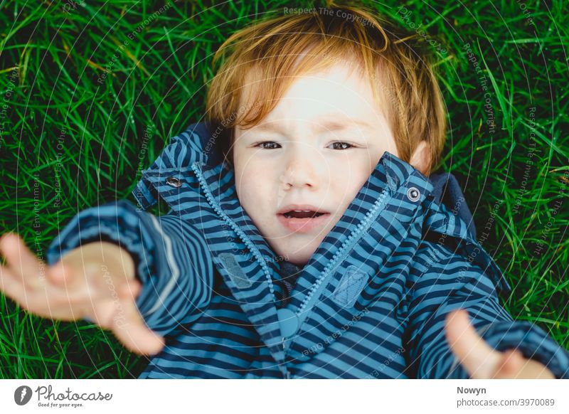 Don't leave me, Mummy! Young boy lying on the ground looking up with outstretched arms blue casual closeup cool face ginger gjeans gras head shot headshot