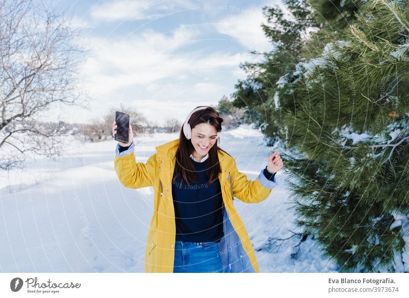 young beautiful woman listening to music on mobile phone and headset in the snow. wearing yellow coat, winter lifestyle mountain technology daytime caucasian