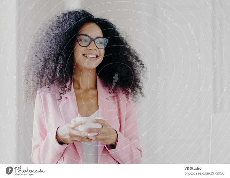 Photo of good looking African American prosperous busineswoman waits for partner in office, drinks coffee discusses future plans with colleague looks away with cheerful expression wears formal clothes