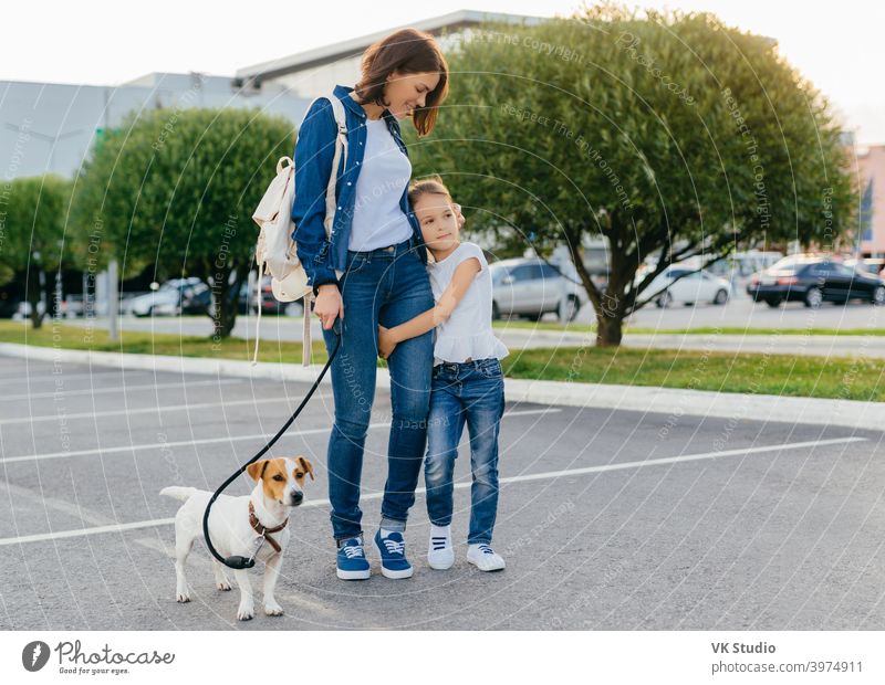 Affectionate mother embraces small daughter, have outdoor stroll together with their favourite pet, enjoy good day and weather, pose against city background. Family, urban style and day off concept