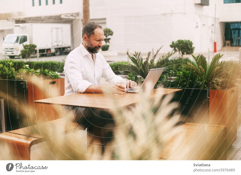 Adult male businessman sitting on the street in a cafe, drinking coffee and working on a laptop millennial freelancer smartphone meetings brainstorming