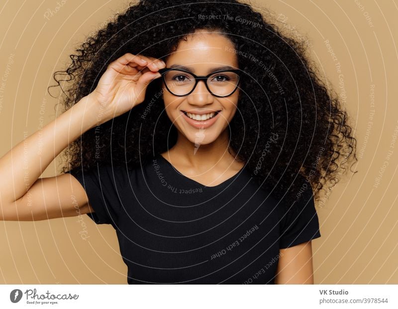 Lovely young African American woman grins at camera, keeps hand on rim of spectacles, glad to hear good news, dressed in casual black t shirt, isolated over brown background. Happiness concept