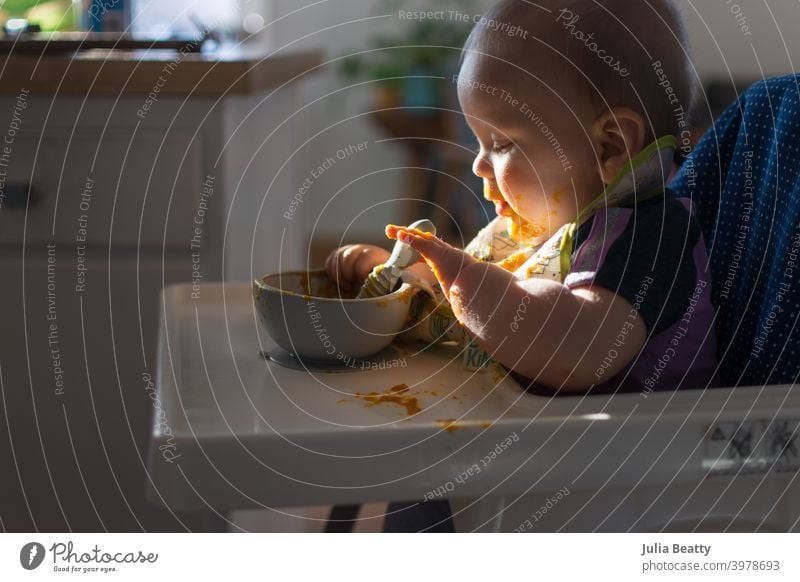 Baby seated in high chair reaching for spoon; dramatic natural lighting and messy tray table baby led weaning first foods finger foods dramatic lighting