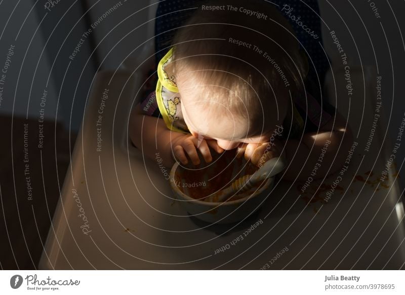 Baby chewing on silicone bowl while seated in high chair; dramatic natural lighting baby led weaning first foods finger foods dramatic lighting spotlight