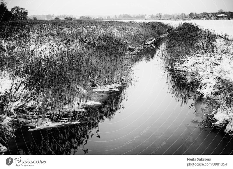 View of the river in early winter at sunset with dry tall grass and the first snow in the foreground / landscape from a bridge over fields in Latvia Autumn