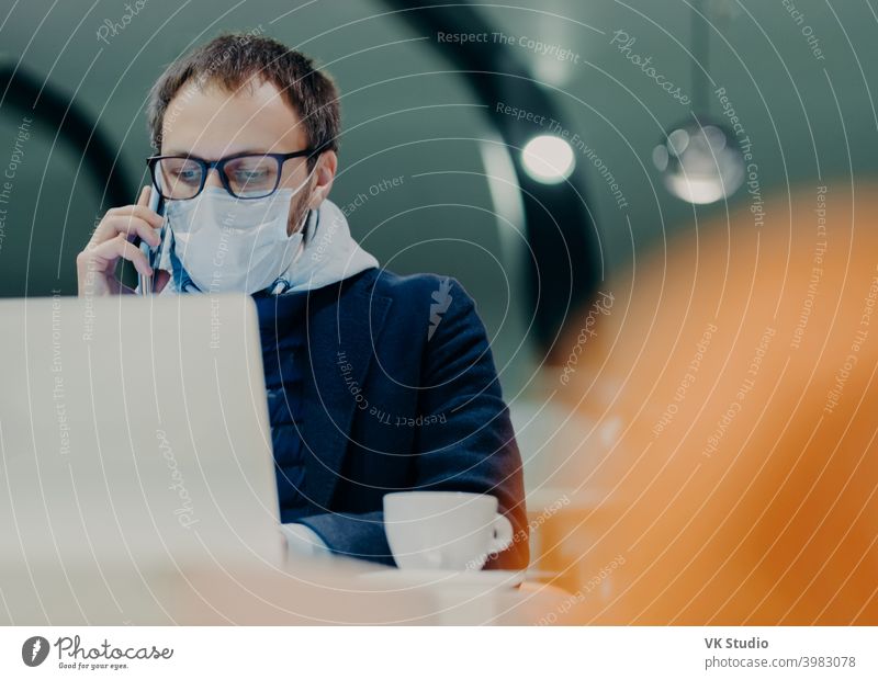 Businessman works remotely from cafeteria, concentrated at display of laptop computer, has telephone talk, wears medical mask on face as protection against flu or coronavirus epidemy. Smart working