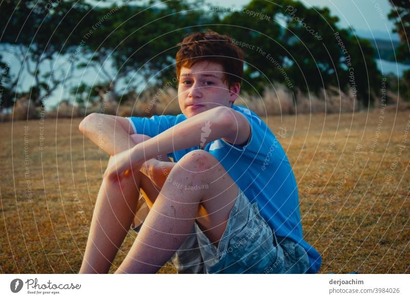 A boy sits with folded arms on the ground. Bushes and shimmering blue water can be seen in the background. He looks into the camera. Young man portrait