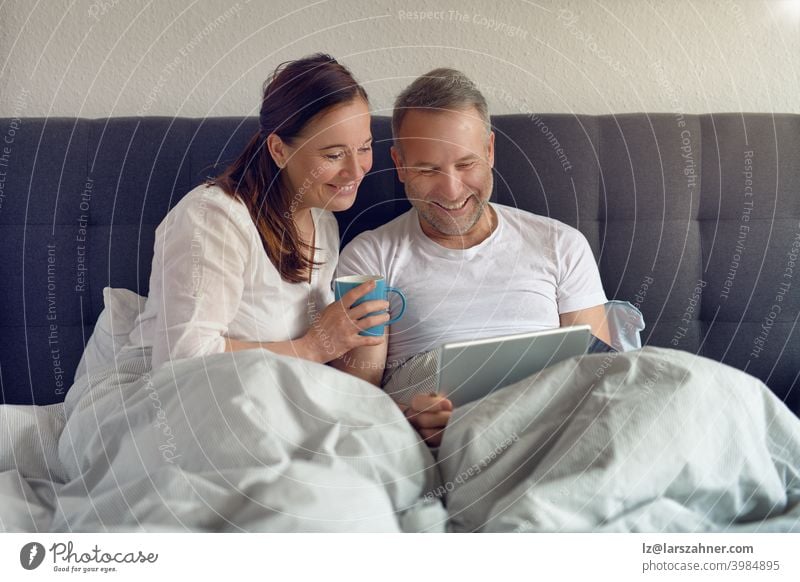 Happy middle-aged couple sitting in bed in the morning drinking coffee and doing online shopping with a digital tablet sofa furniture family room reclining