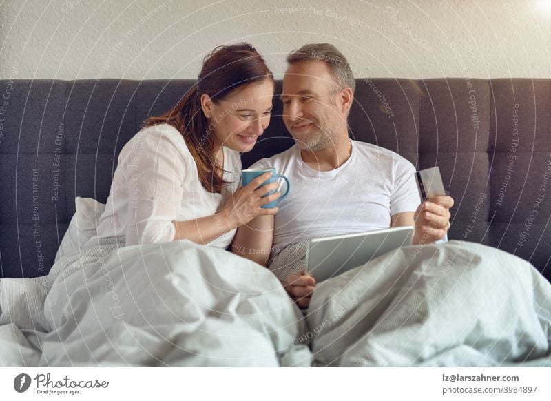 Happy middle-aged couple sitting in bed in the morning drinking coffee and doing online shopping with a digital tablet sofa furniture family room reclining