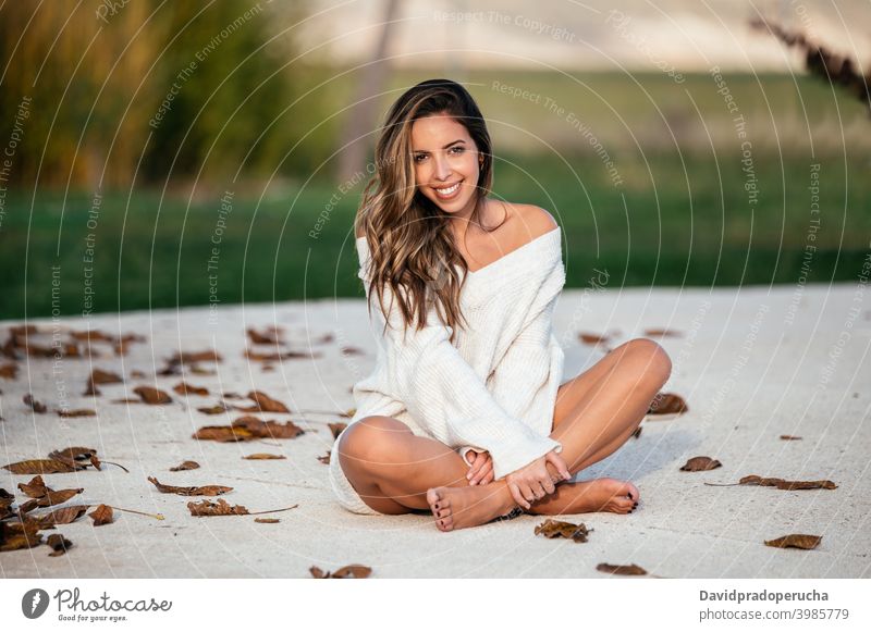 Delighted woman resting in yard on autumn day weekend smile barefoot legs crossed cement ground happy cheerful joy relax daytime leaf dried fall long hair glad