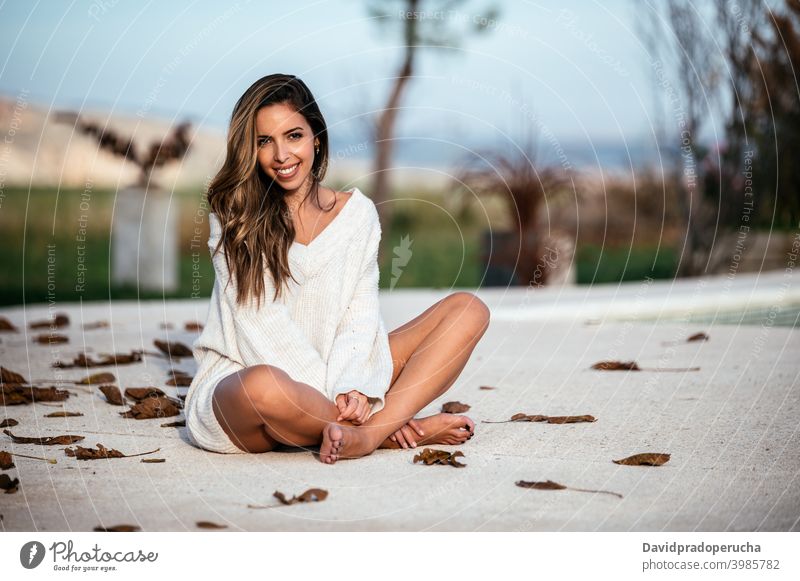 Delighted woman resting in yard on autumn day weekend smile barefoot legs crossed cement ground happy cheerful joy relax daytime leaf dried fall long hair glad