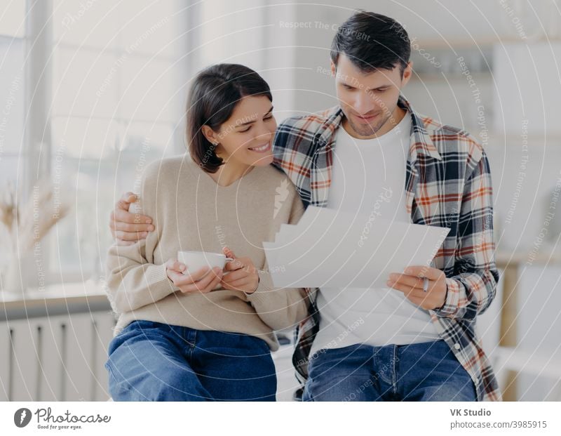 Caring man embraces his wife, look through documents with happy expressions, study payment bills, drinks coffee, dressed in casual wear, pose in spacious room with blurred background. Family and work