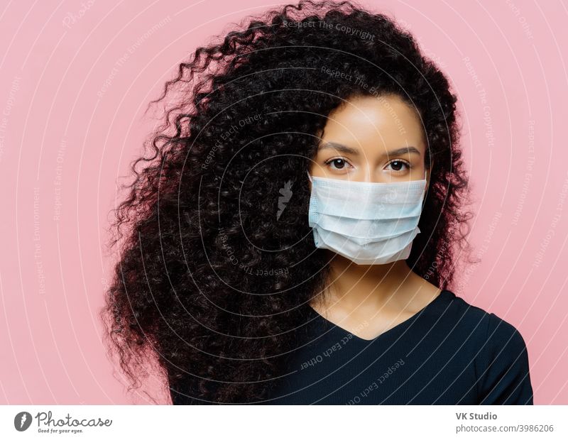 Serious dark skinned young woman being on self isolation at home, wears protective medical mask, being on quarantine at home, dressed casually, poses against pink background. Coronavirus disease