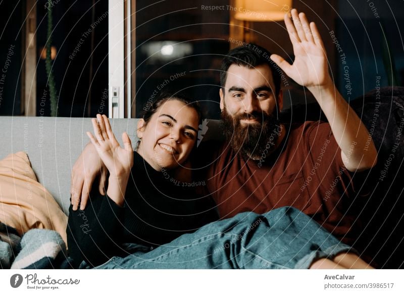 A young couple on the couch greeting to the camera and smiling while on pandemic affectionate embracing hugging humor wife husband photograph ethnicity living