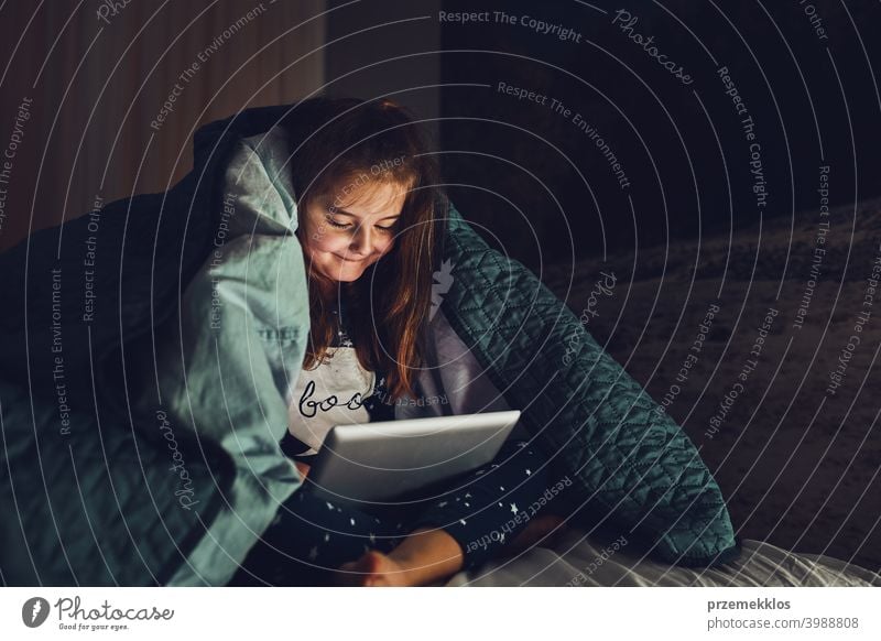 Little girl having fun watching, playing and listening to stories on tablet computer laying under duvet night bedtime child home bedroom read kid relaxation