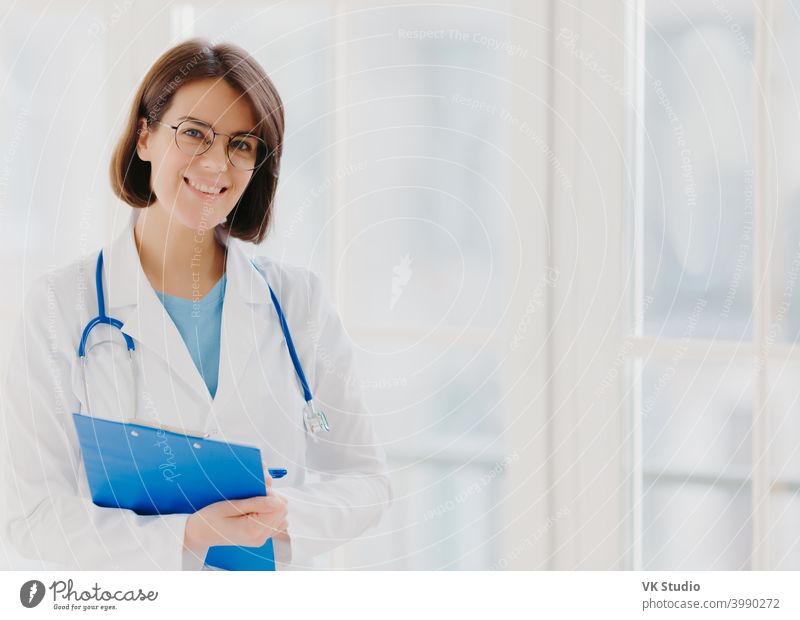 Good looking female expert creats medical prescription, writes down information in clipboard, thinks about good advice for patient, stands indoor, copy space for your information. Medicine concept