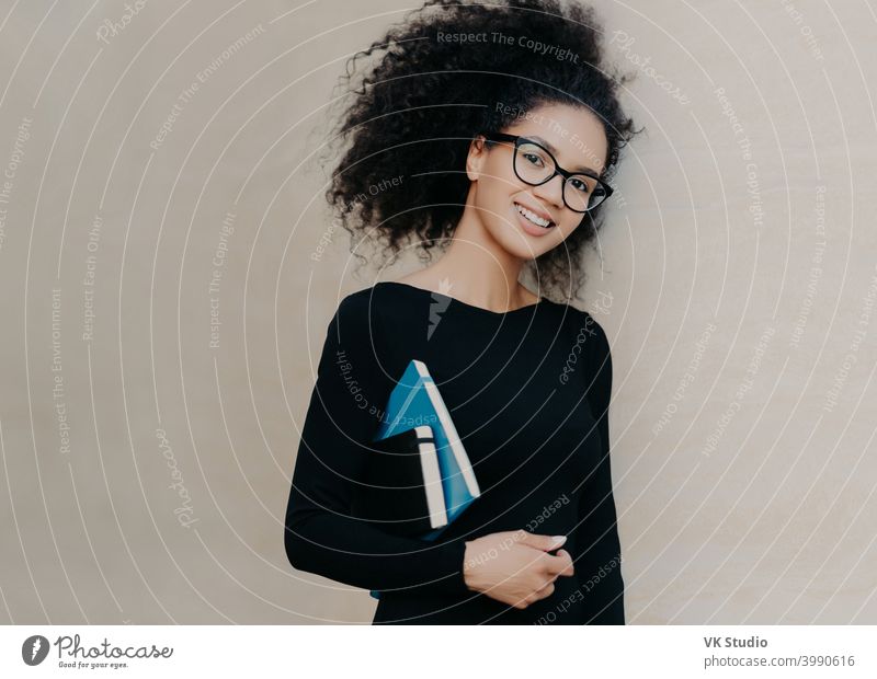 Positive curly African American student carries notepad and diary, wears black clothes, transparent glasses, enjoys studying in university, isolated on grey background. Cute woman with notebook