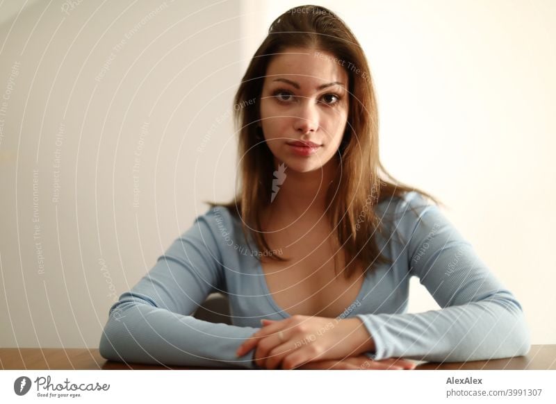 Portrait of a young brunette woman with a light blue top in a bright room at a wooden table Young woman pretty Brunette Long-haired Slim Feminine teen