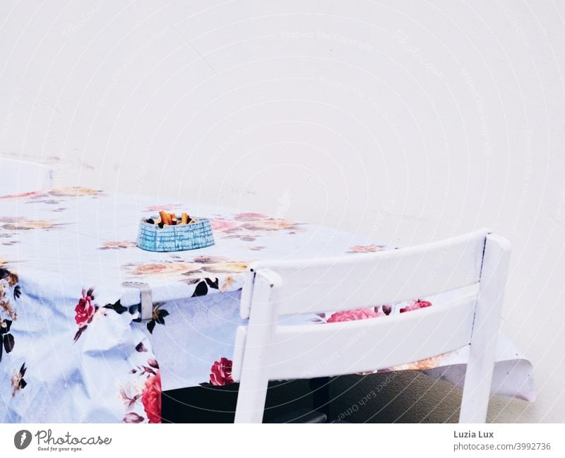 Smoker's corner or an overflowing blue ashtray on a table with an old-fashioned waxed blanket, floral pattern and summery white Ashtray smoking corner