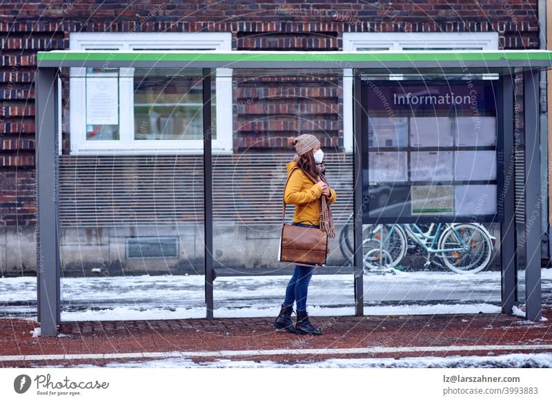 Midde-aged brunette woman standing at a bus stop, wearing a protective face mask due to corona virus, waiting for her bus to bring her to work middle-aged