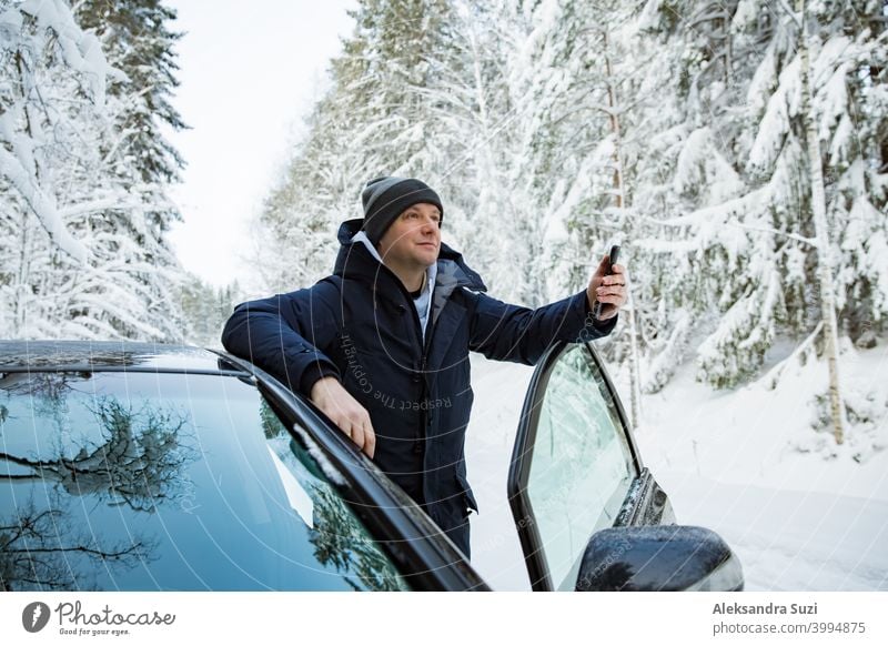 Man in warm winter clothes standing at the car, using phone. Snowy winter country road, car covered with ice, Beautiful forest under the snow. help call