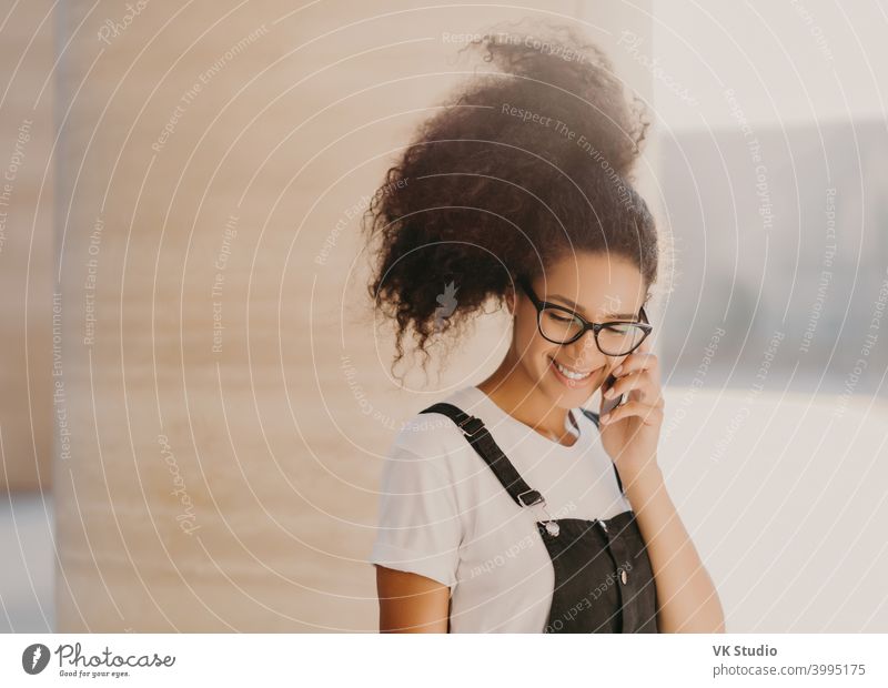 Millennial Afro woman speaks via cell phone, uses roaming connection, looks down, enjoys pleasant conversation, wears white t shirt and overalls, rests outdoors, calls via app, has good mood