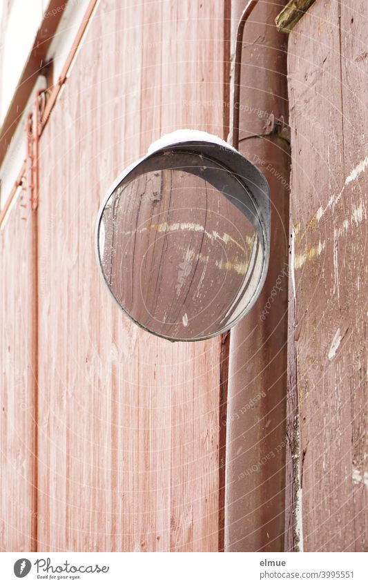 A round, snow-covered traffic mirror is mounted next to a large, reddish-brown wooden gate, reflecting the two horizontal scratches on the gate. traffic mirrors