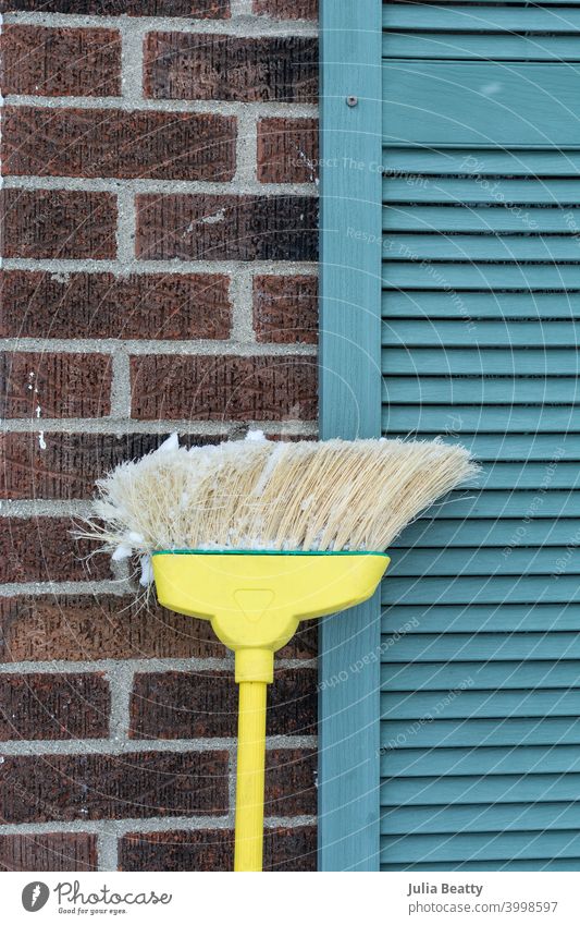 Broom covered in snow leaning against brick home with blue shutters; cleanup after a snow storm broom brush isolated cleaning flurries cold winter accumulation