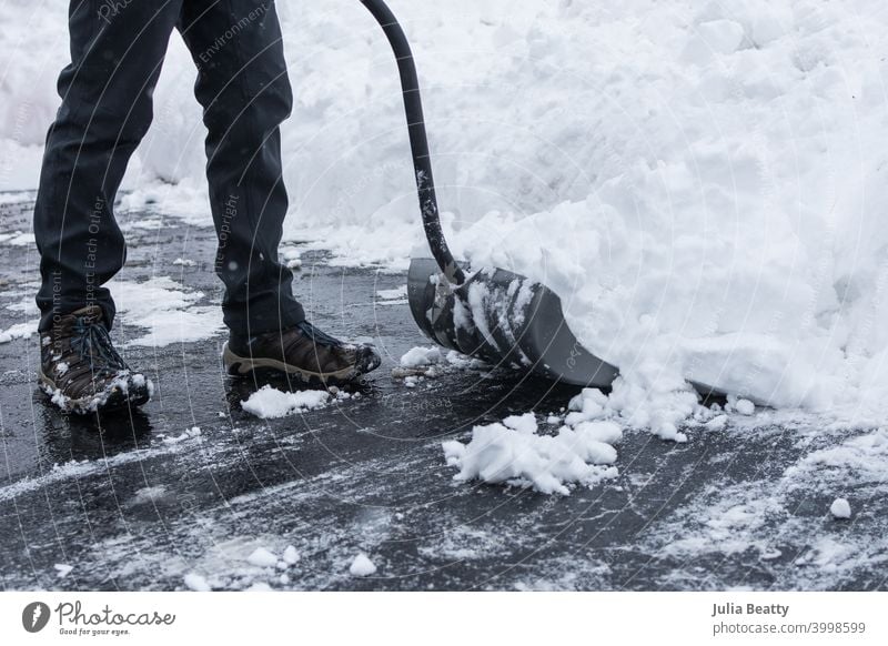 Snow being shoveled off a black asfalt driveway with shovel; person wearing gray pants and boots; big pile of snow snowstorm clear path home owner man help