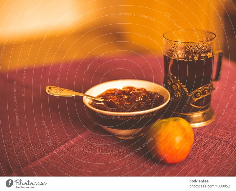 a still life with a tea glass in a metal saucer, next to it a porcelain bowl with apple jelly, in which a silver teaspoon is stuck, in front of both lies an apple. Everything stands on a rust-red tablecloth. Blurred foreground and background