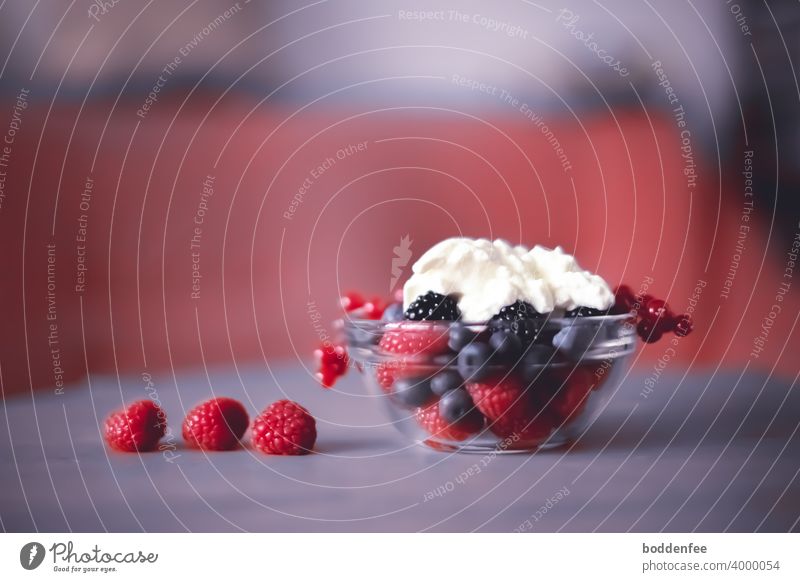 a glass bowl with fresh raspberries, blackberries blueberries and currants, on top in the <cup ripened yogurt. Three raspberries lie on the soft purple tablecloth. Blurred foreground and background