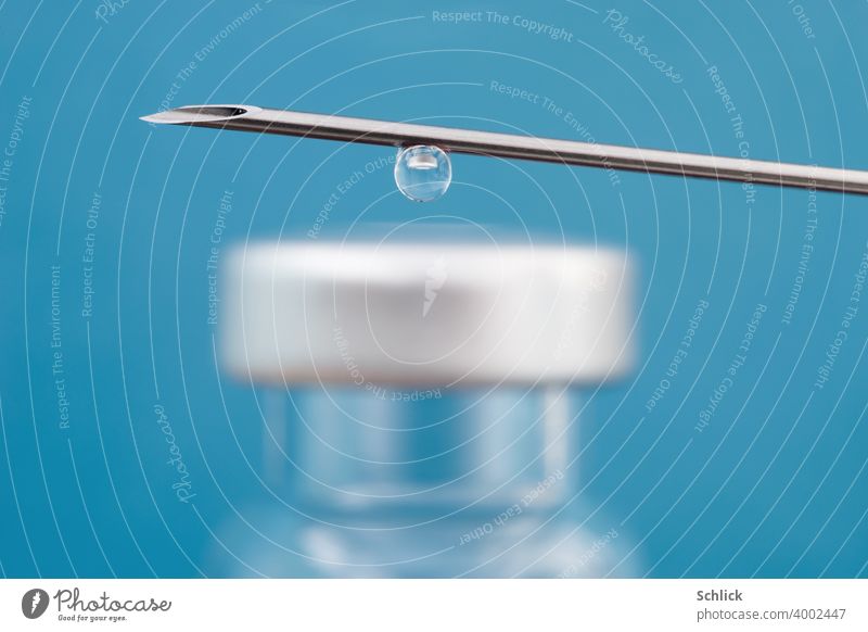 A drop of a vaccine hangs from a cannula and shows the reduced image of a small glass bottle with septum macro with shallow depth of field and blue background