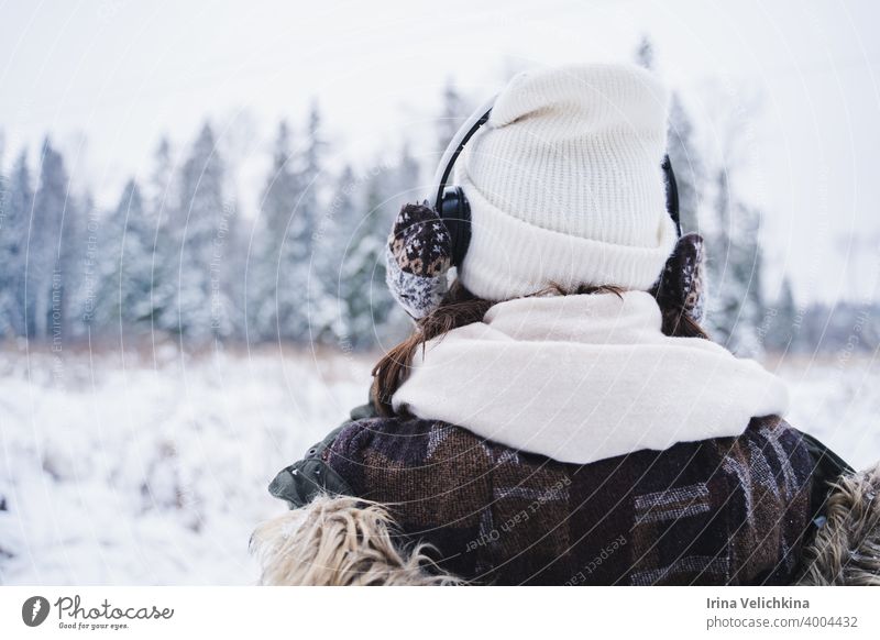 Young girl, woman in knitted mittens listening to music on headphones. Walking in beautiful winter forest among trees, firs, covered with snow. Magnificent nature. Fashionable image,clothes,parka,hat