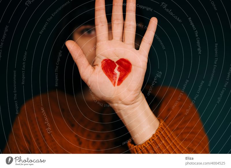 A woman holds her hand, on which a broken red heart is painted, in front of her face. Concept heartbreak. Heart shattered Lovesickness relational Emotions Red