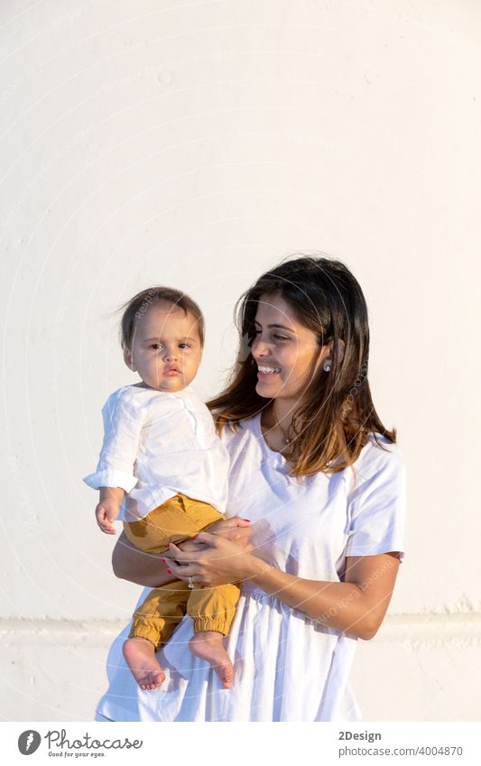 Young mother holding her little baby against white wall in sunny day child newborn love family woman care motherhood young childhood cute person happy girl