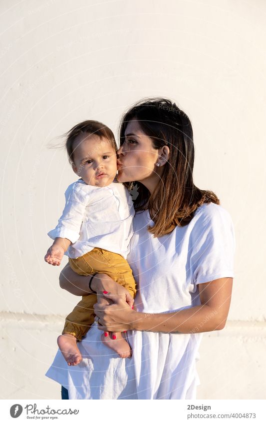 Young mother holding her little baby against white wall in sunny day child newborn love family woman care motherhood young childhood cute person happy girl