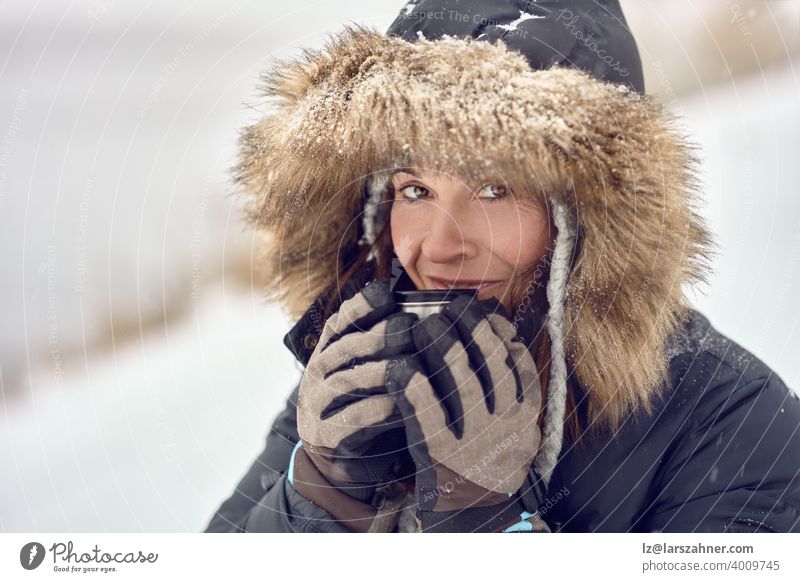 Happy smiling woman wearing a fur trimmed hooded jacket enjoying a mug of coffee cradled in her gloved hands outdoors in winter snow in a concept of the seasons in a close up portrait