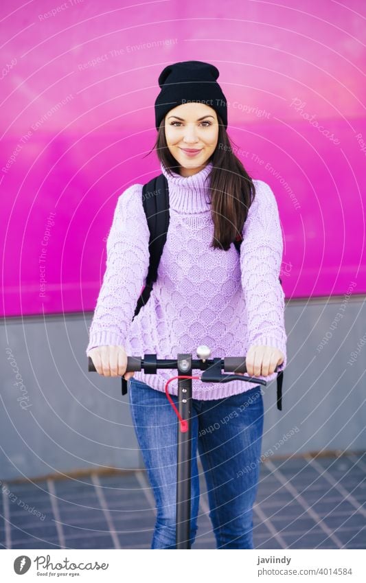 Young woman in her twenties riding an electric scooter. e-scooter girl winter cap backpack sweater urban vehicle street caucasian technology ride lifestyle 20s