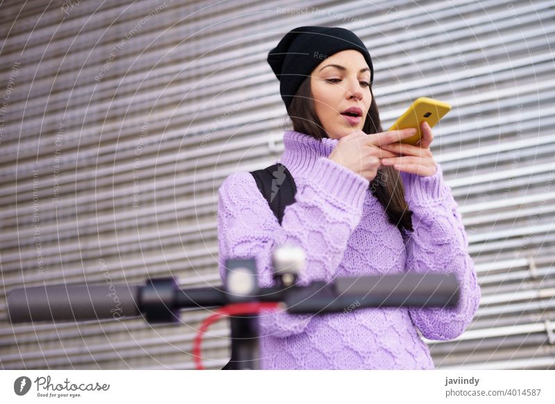 Woman in her twenties with electric scooter recording voice note with a smartphone outdoors. woman e-scooter device girl winter cap backpack urban street