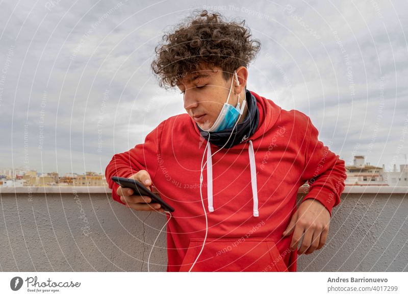 Young attractive man with curly hair dressed in a red sweatshirt using the mobile on the terrace of the house wearing a mask to protect himself from coronavirus