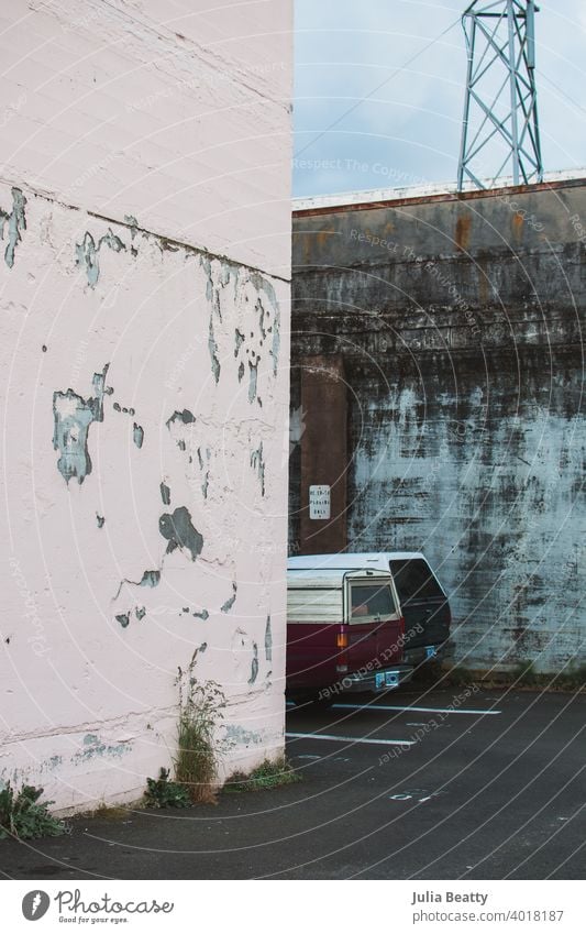 Parking lot with two old trucks, chipped pink paint on cement wall, weeds, and tower; an industrial and worn Car flatbed canopy shell truck topper antique aged