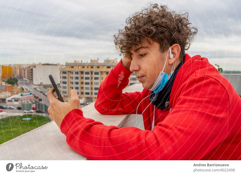 Young attractive man with curly hair dressed in a red sweatshirt using mobile in living room at home wearing a mask to protect himself from coronavirus