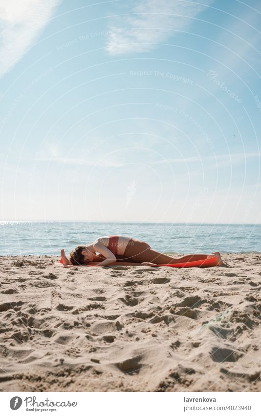 Young female in orange top and leggins practising Yoga split variation (Hanumanasana) on a beach in the morning woman yoga fit training splits sporty leggings