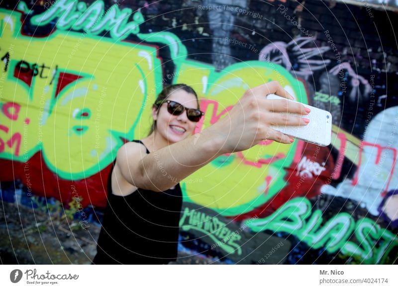 Young woman taking a selfie in front of a graffiti wall smartphone Selfie vanity youthful Self portrait Face Telephone Cellphone mobile phone camera pose