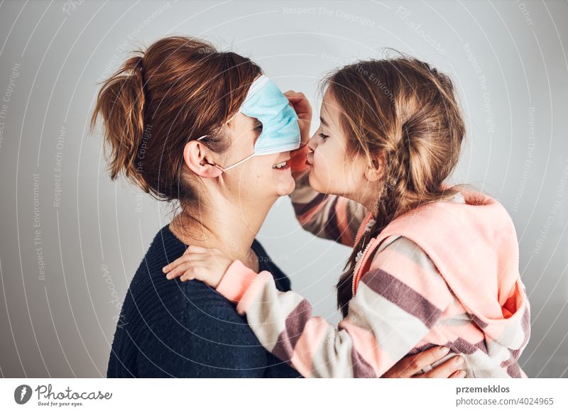 Little girl moving her mom's face mask on eyes and giving a kiss her mother. Funny moments during covid-19 pandemic affection affectionate bond care caucasian