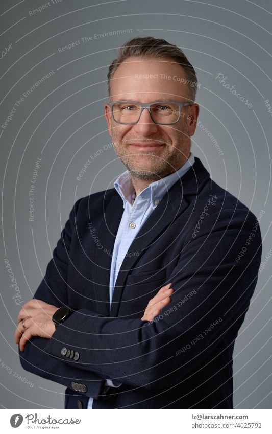 Smiling mature businessman in glasses, wearing suit without a tie, standing against grey background and posing at camera with his arms folded. Half-turn half-length studio portrait