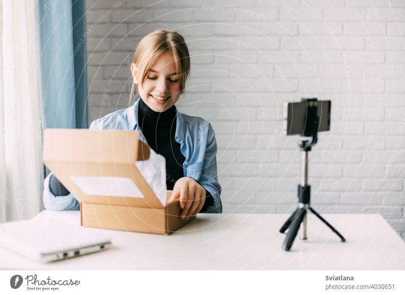 A teen blogger records a video and unpacks a box for his blog on a smartphone on a tripod at home. Communication of a video blogger girl on social networks with subscribers and friends