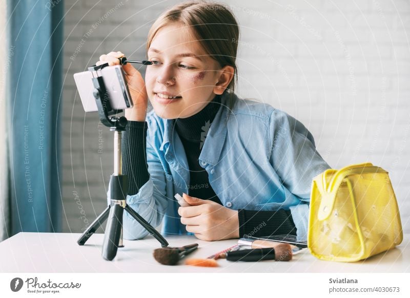 A teenage girl shows how to dye her eyebrows and eyelashes while recording a video for her blog on a smartphone on a tripod at home. friends blogger camera