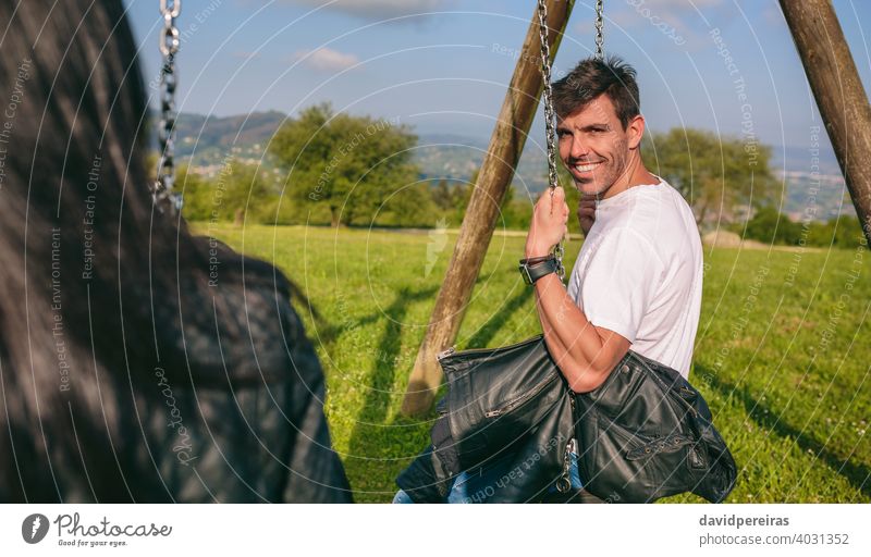 Happy young man looking camera sitting on a swing attractive smile happy recreational area girlfriend friendly retro handsome guy fashion pretty t-shirt park